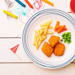 Small kid's meal - chicken nuggets, french fries, carrot and green peas. Colorful dinner on white wooden table. Plate captured from above (top view, flat lay). Layout with free copy (text) space.